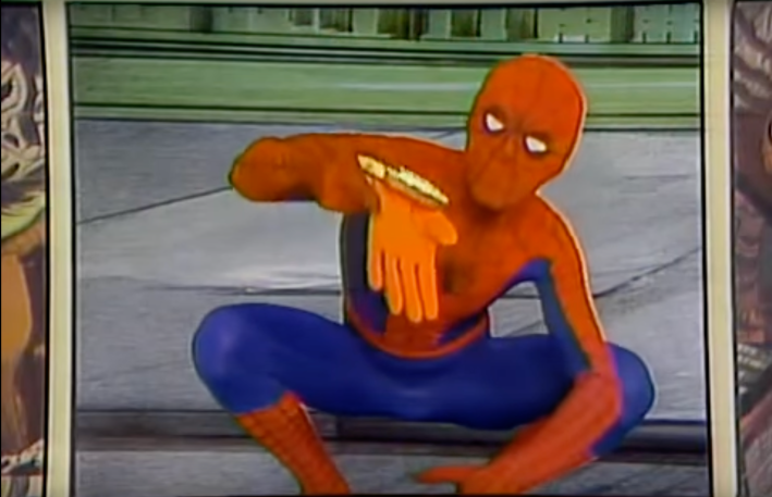 An actor as Spider-Man, in his red and blue tights, sits on a curb, holding a sandwich that has rubber gloves sticking out of it.